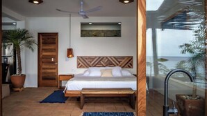 Ein Schlafzimmer mit Bett, Holz-Kopfteil, Deckenventilator und Blick auf das Meer durch eine Glastür.