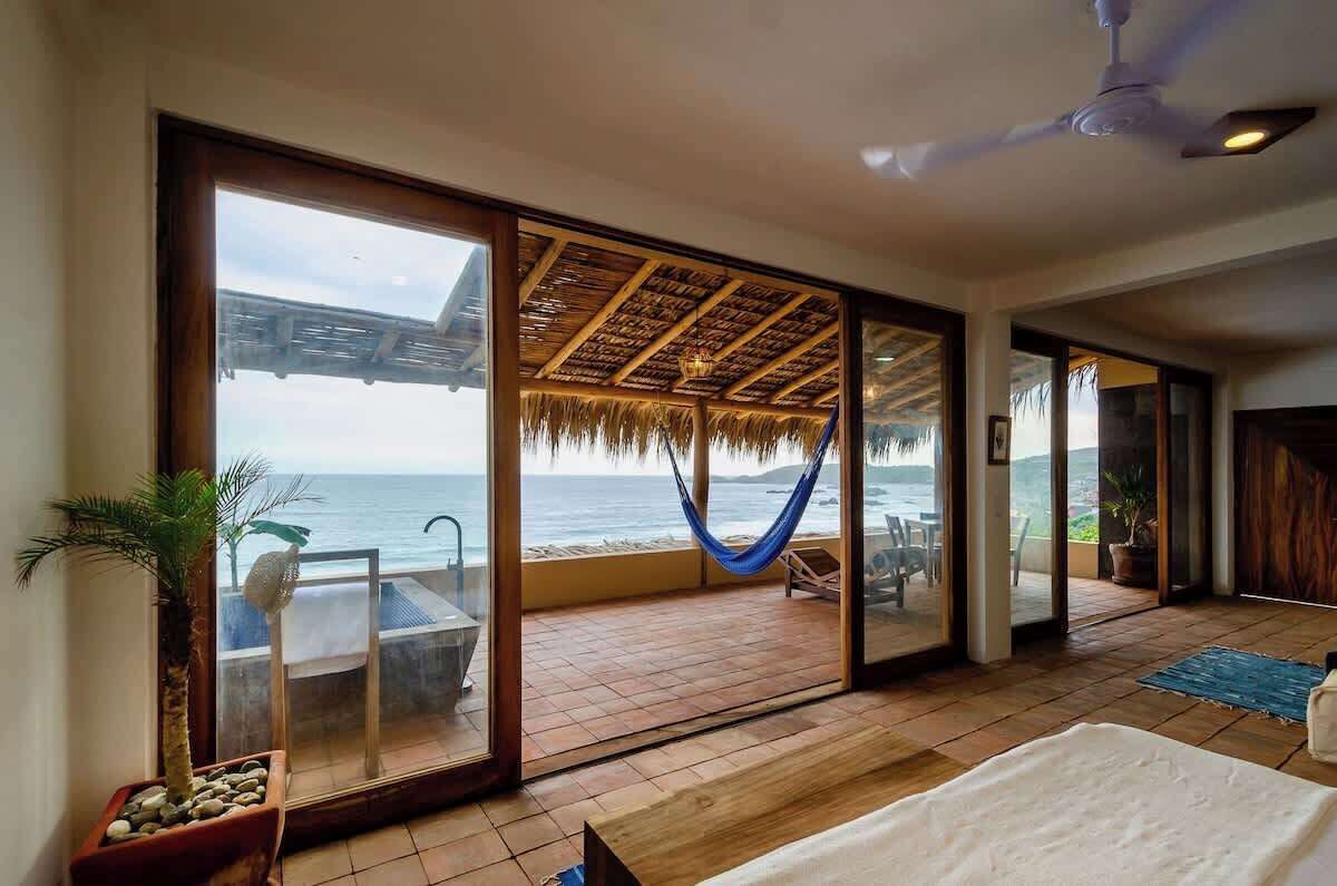 Una habitación con un gran ventanal corredizo que da a un balcón con vista al océano. En el balcón hay una hamaca, un ventilador de techo y una cama con cubrecama blanca.