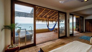 Ein Zimmer mit einer großen Glasschiebetür, die auf einen Balkon mit Meerblick führt. Auf dem Balkon befindet sich eine Hängematte, ein Deckenventilator und ein Bett mit weißer Bettdecke.