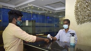 A reception desk with a glass partition, a blue curtain in the background, and a decorative wall piece. - Hotel Pondichery (Puducherry)