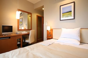 Habitación de hotel con una cama, un escritorio con computadora y teléfono, una silla, una lámpara de noche y un cuadro en la pared.