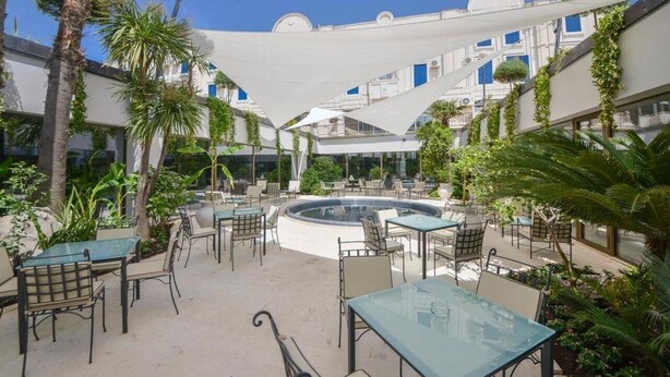 Un patio al aire libre con mesas y sillas, rodeado de follaje y una fuente central.