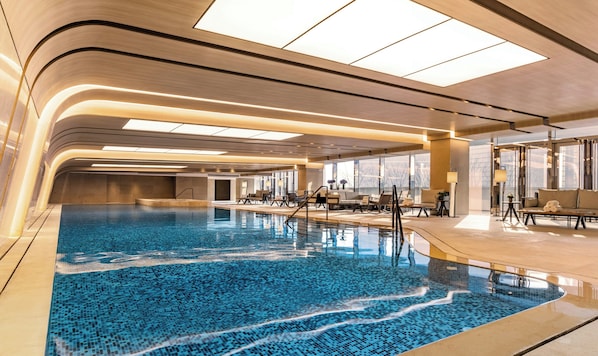 A modern indoor swimming pool with a clear blue mosaic floor, surrounded by lounge areas with seating and tables, and large windows providing natural light. - Conrad Hangzhou (Hangzhou)