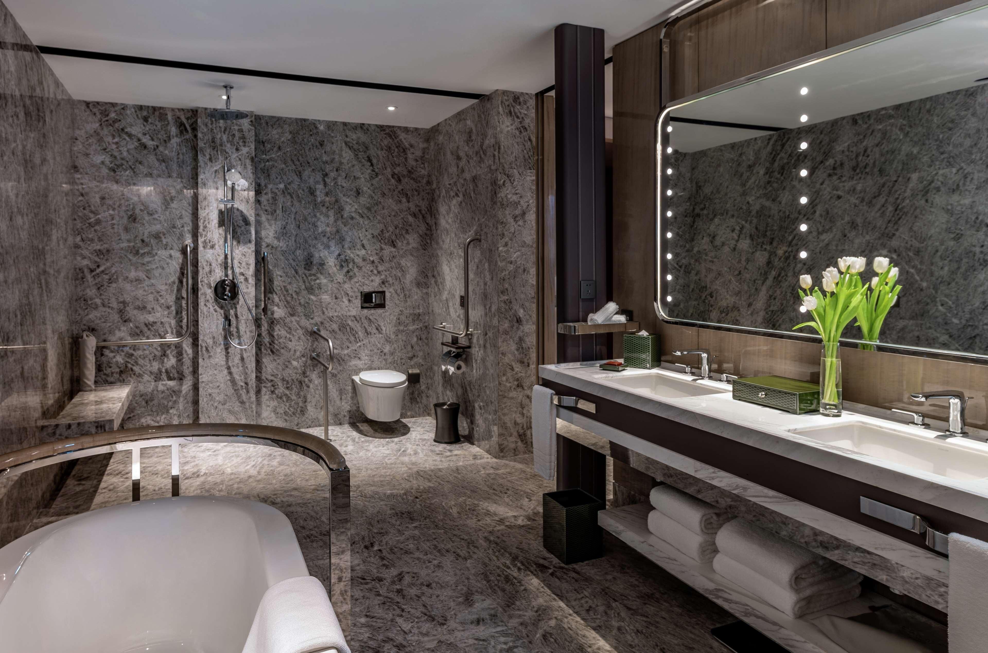 A modern bathroom with a marble shower, a bathtub, a large mirror with lights, and a vanity with a sink.