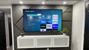 Televisão de tela plana na parede, com design moderno, exibindo a interface Roku TV e diversas opções de streaming.