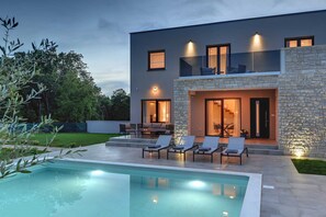 Een modern huis met twee verdiepingen, een zwembad, buiten zitplaatsen en een stenen gevel.