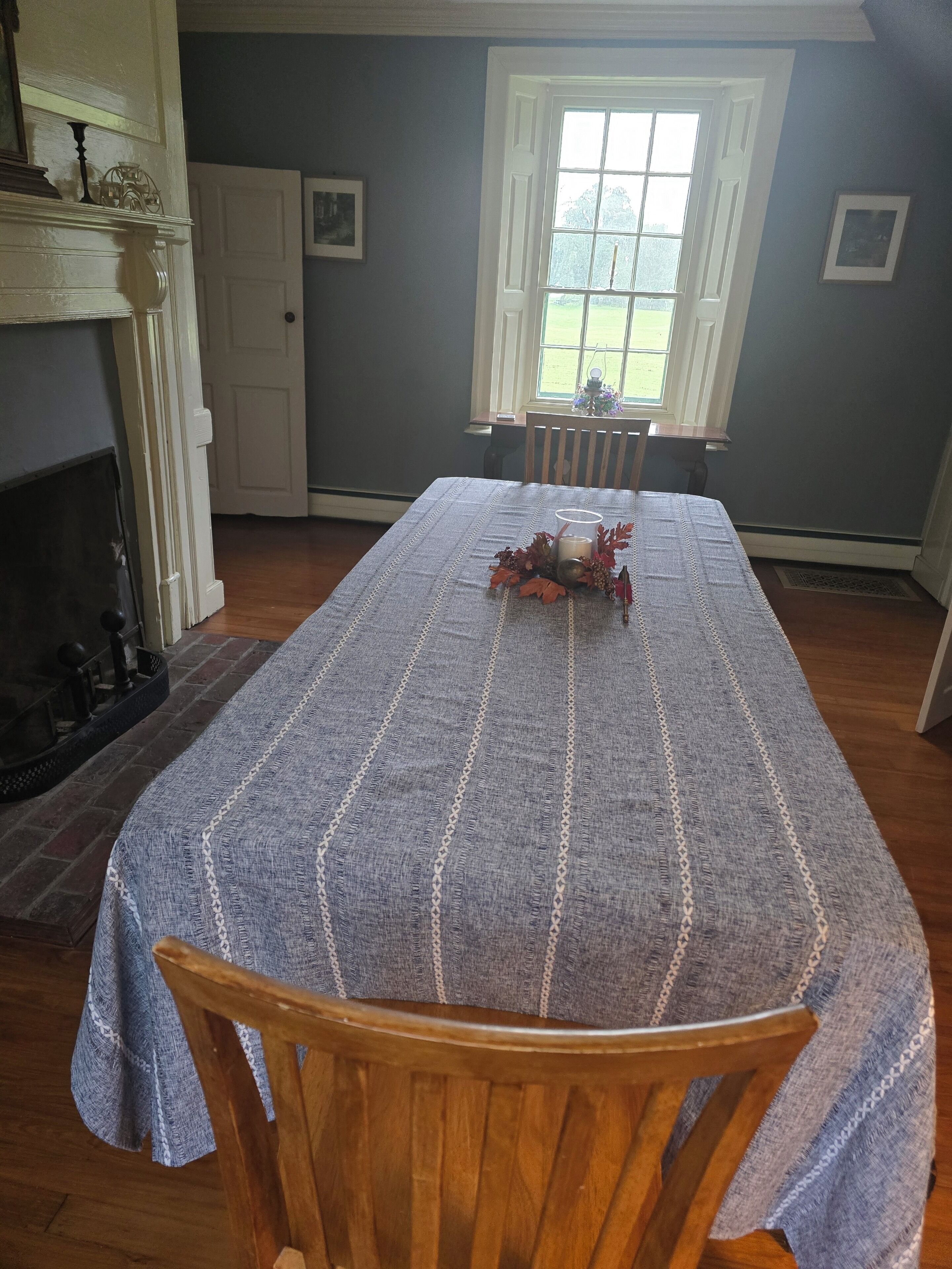 Un comedor con una mesa larga cubierta por un mantel gris, sillas de madera, una chimenea y una ventana con vistas al verdor.