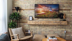 Ein Wohnzimmer mit Backsteinwand, einem an der Wand befestigten Fernseher, einem hölzernen Regal, einem Korbstuhl, einem grauen Sofa und einem runden Couchtisch.