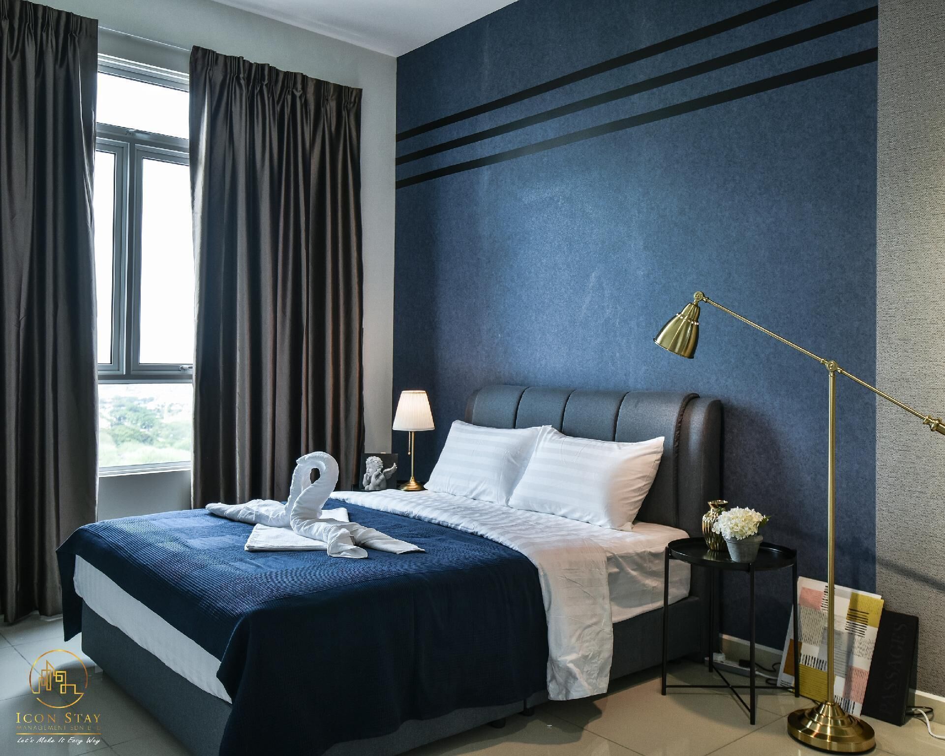 A hotel room with a large bed, a nightstand, a lamp, and a view of the outdoors through a window.