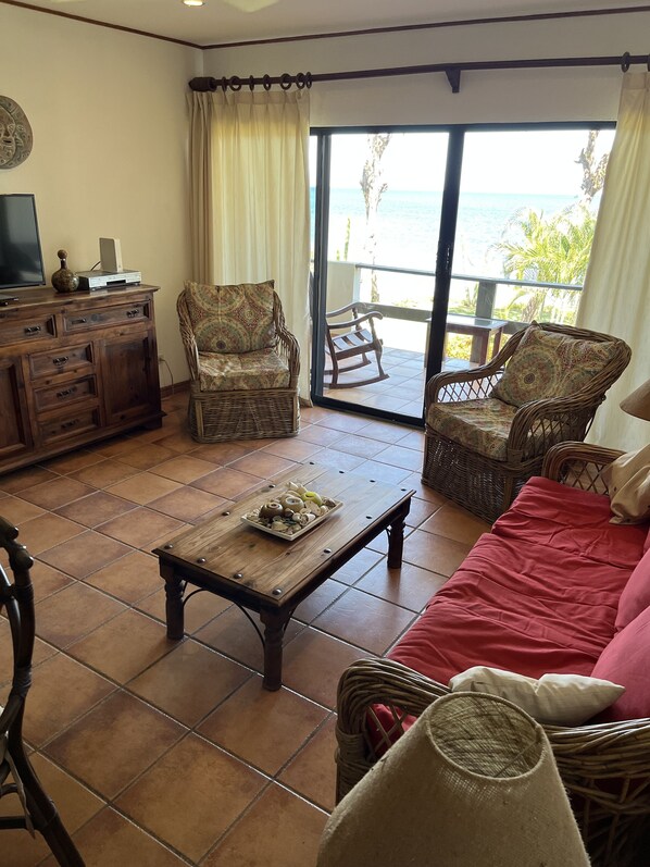 A living room with a wooden coffee table, wicker furniture, a red sofa, and a view of the ocean through large sliding doors. - Marina Cove 610 Beautiful Beachfront Condo! (Playa Flamingo)