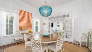 A dining room with a round wooden table surrounded by white chairs, a blue chandelier, and a large window with white curtains.