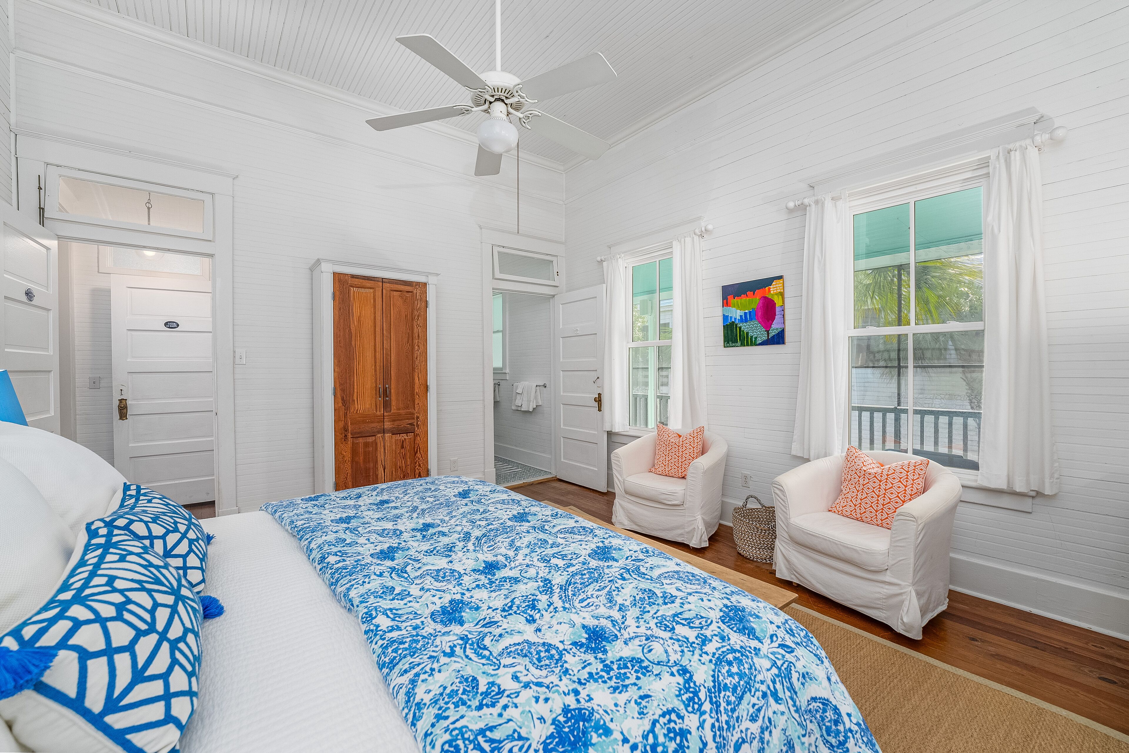 A bedroom with a bed, two chairs, a ceiling fan, and a window with curtains.