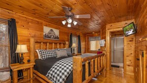 Quarto de cabana de madeira com cama, ventilador de teto, televisão e banheiro com chuveiro.