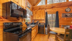 Interior de uma cabana de madeira com cozinha equipada, incluindo fogão, forno, micro-ondas e máquina de lavar louça. Há uma mesa de jantar com cadeiras e uma janela com cortinas.