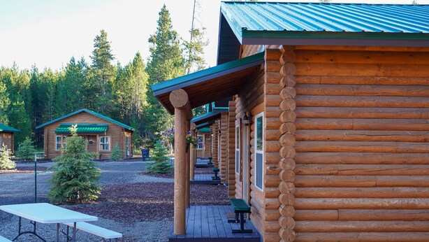 A log cabin with a porch, outdoor seating, and a small tree in front. - Cabin Village (Island Park)