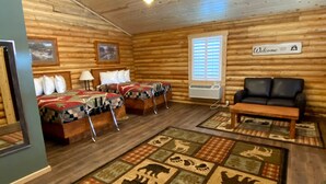 A log cabin-style room with a bed, two chairs, a sofa, a rug with animal prints, a chandelier, and a 'Welcome' sign. - Cabin Village (Island Park)