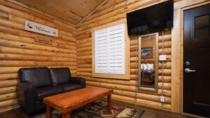 A log cabin interior with a leather sofa, a wooden table, a welcome sign, a window with blinds, and a door with a mirror. - Cabin Village (Island Park)