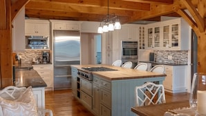 A spacious kitchen with wooden beams, white cabinetry, a central island, and a dining area with white chairs. - Rally Point I Soaring Timber Frame Estate I Beaches, Trails, Newport (Middletown)
