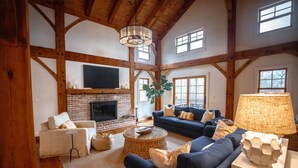 A spacious living room with a brick fireplace, a large TV mounted on the wall, and a cozy seating area with a blue sofa and a white armchair. - Rally Point I Soaring Timber Frame Estate I Beaches, Trails, Newport (Middletown)