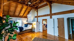 A spacious room with a high ceiling and exposed wooden beams, a large bed with a headboard, and a view of another room through an open door. - Rally Point I Soaring Timber Frame Estate I Beaches, Trails, Newport (Middletown)
