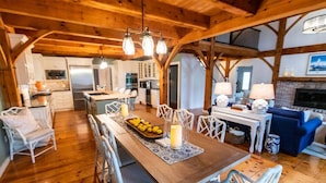 A spacious kitchen with wooden beams, a dining area with a wooden table and chairs, and a living area with a sofa and fireplace. - Rally Point I Soaring Timber Frame Estate I Beaches, Trails, Newport (Middletown)