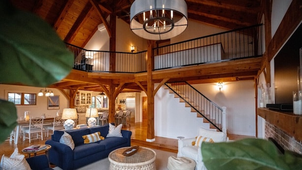 A spacious living room with a high ceiling, wooden beams, a staircase, and a dining area. - Rally Point I Soaring Timber Frame Estate I Beaches, Trails, Newport (Middletown)