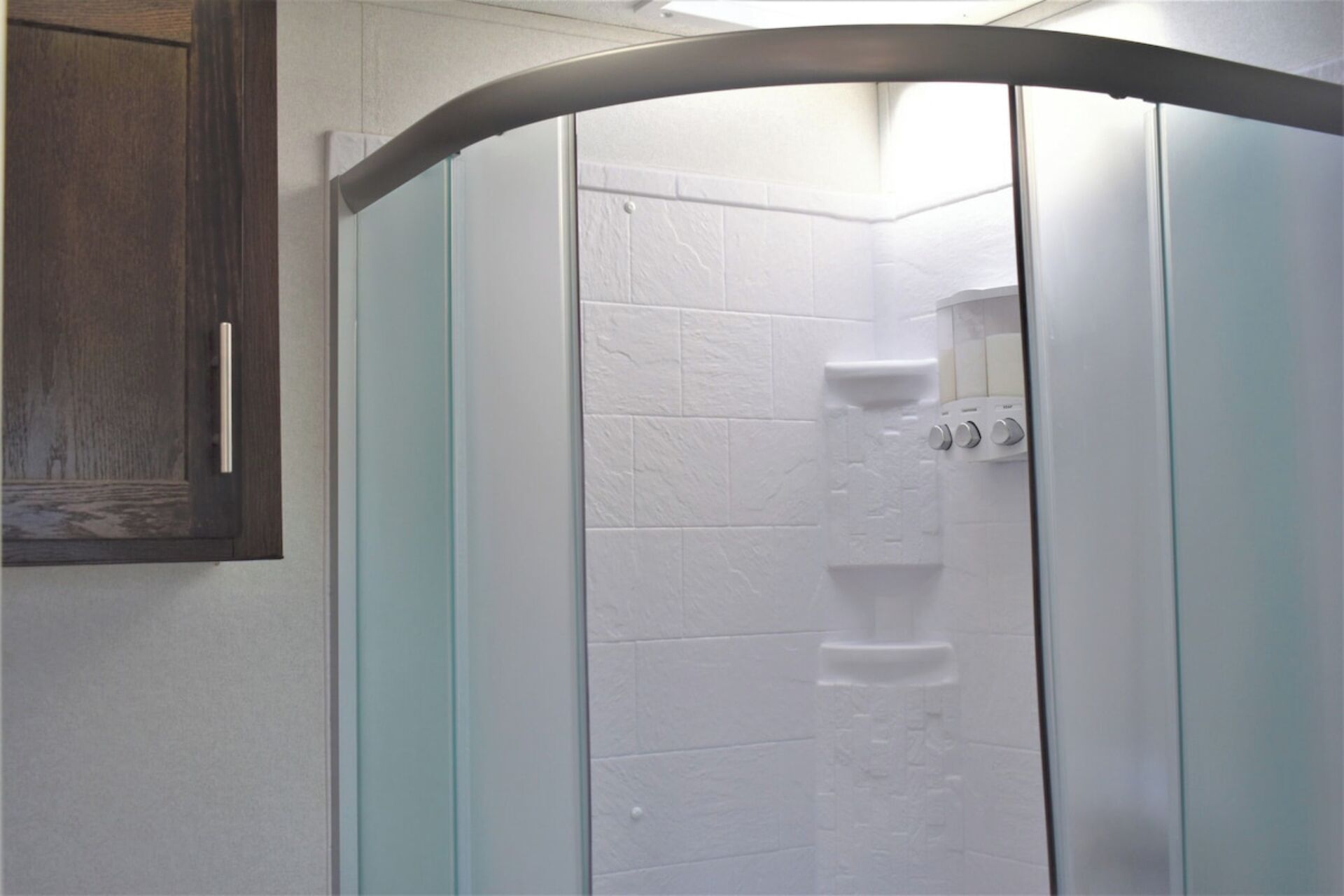 A shower with a curved glass door, white tiles, and a wooden cabinet to the left.