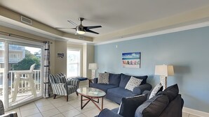 A living room with a blue sofa, a patterned armchair, a glass coffee table, and a ceiling fan. - Gull Reef 614 |  Spacious, Pool Access, Walk to Beach (Tybee Island)