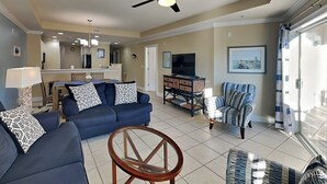 A living room with a blue sofa, a round coffee table, and a patterned armchair. There is a television on a stand and a ceiling fan above. - Gull Reef 614 |  Spacious, Pool Access, Walk to Beach (Tybee Island)