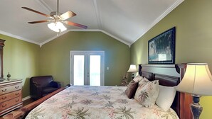 Een slaapkamer met een bed, een plafondventilator, een nachtkastje met een lamp, een dressoir, een stoel en een deur die leidt naar een balkon.