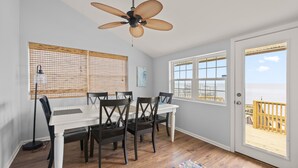 A dining area with a white table and dark chairs, a ceiling fan, and a view of the beach through a door.