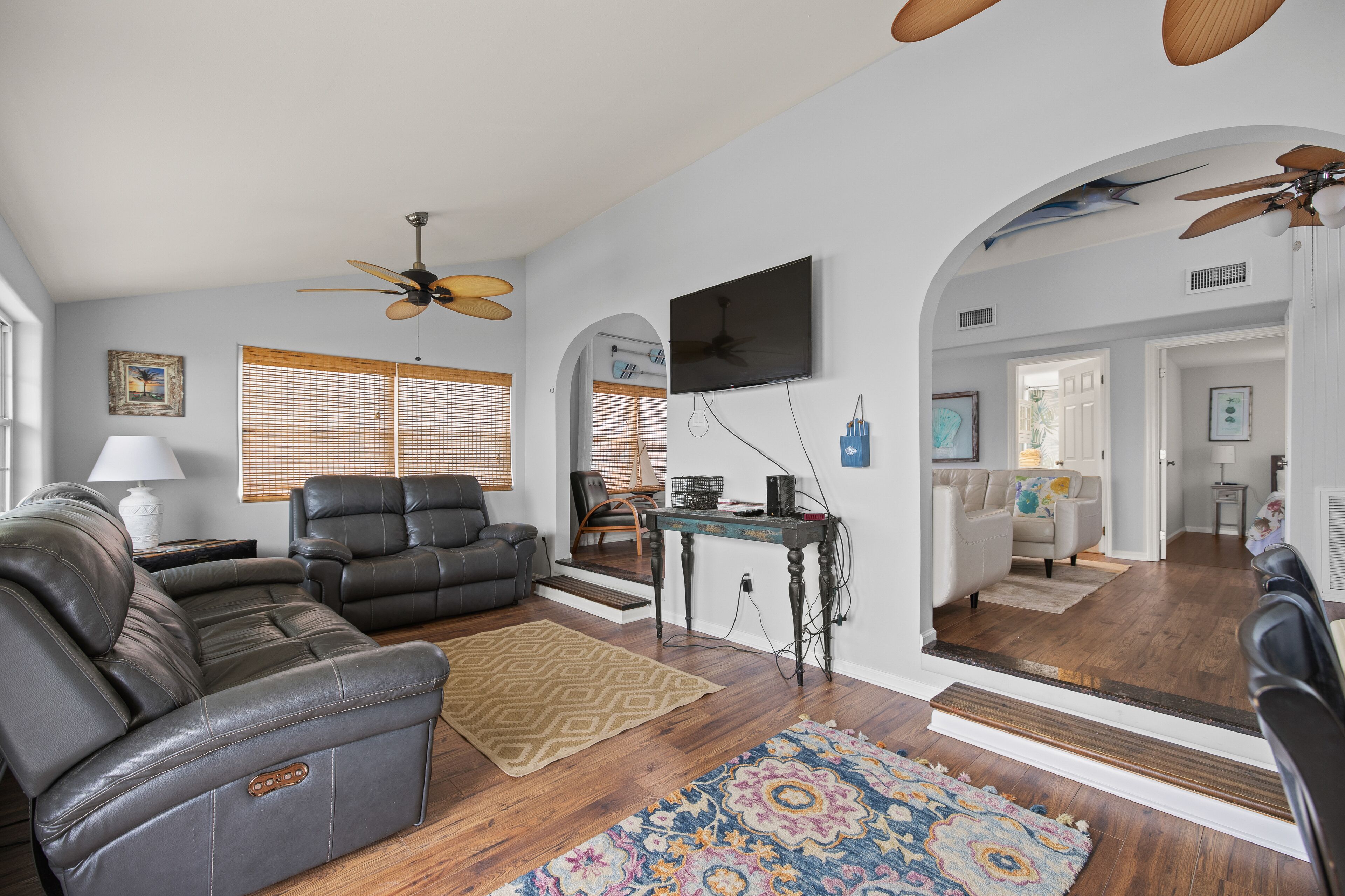 A living room with a sectional sofa, a coffee table, and a TV mounted on the wall. There are wooden floors, a patterned rug, and a ceiling fan.