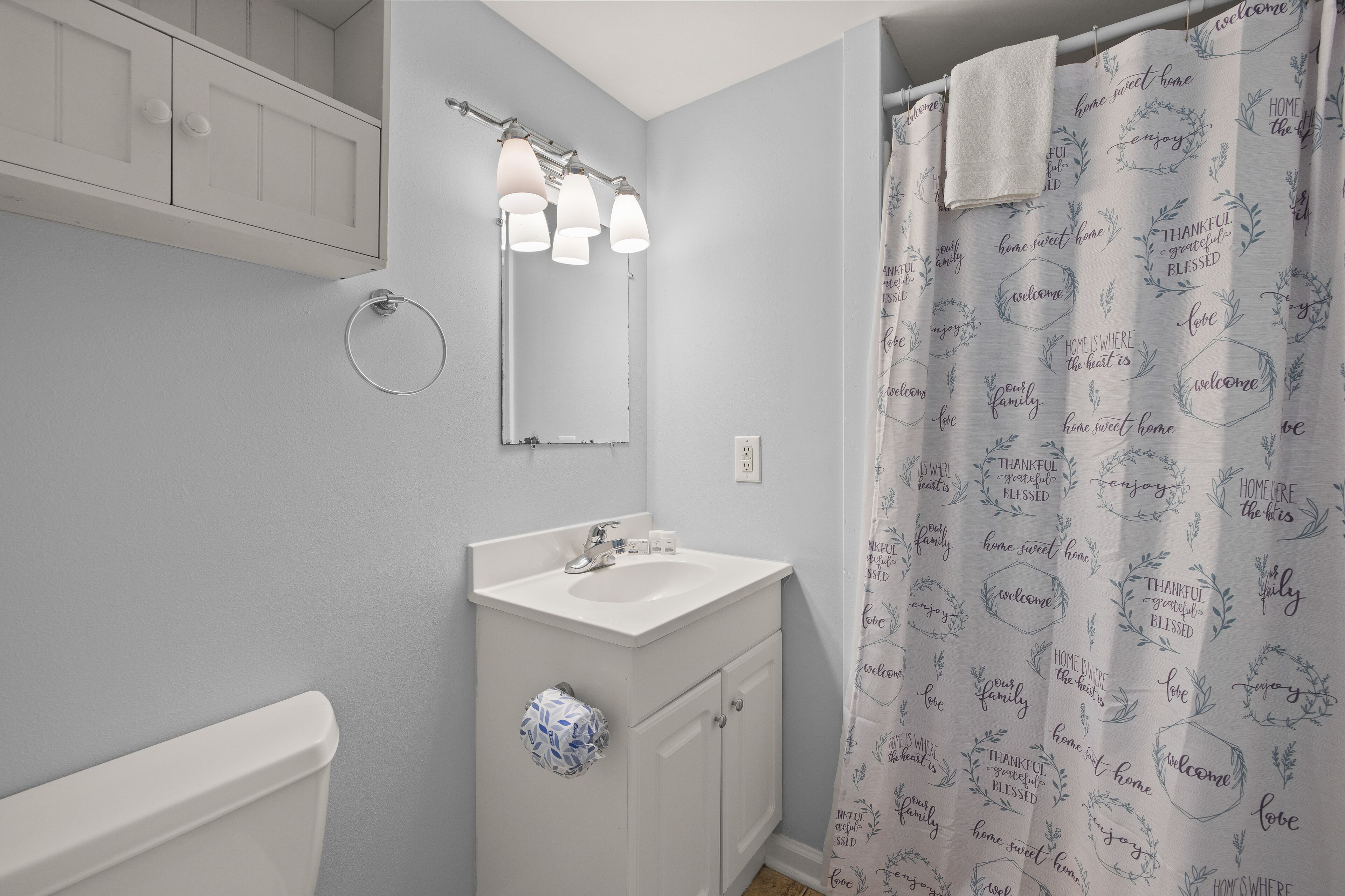 A bathroom with a white sink, mirror, and cabinet, a towel hanging on the wall, and a shower curtain with positive phrases.