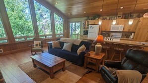 A cozy living room with a wooden floor, a sofa, a coffee table, and a fireplace. There is a kitchen area with wooden cabinets and a refrigerator. - Hanging Rock Get Away (Bigfork)