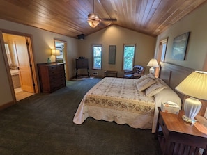 A bedroom with a large bed, a dresser, a chair, and a desk with a lamp. There are windows and a ceiling fan.
