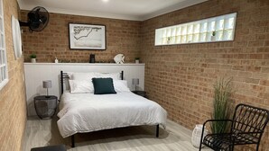 Een slaapkamer met een bakstenen muur, een bed met wit en gestreept beddengoed, een zwarte stoel en een raam met gordijnen.