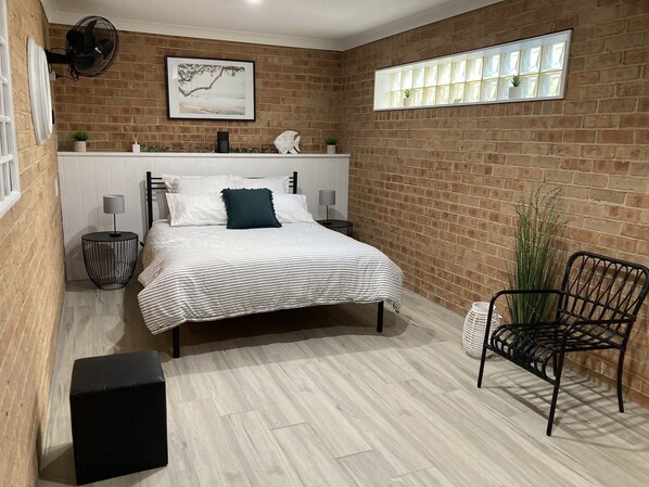 A bedroom with a brick wall, a bed with white and striped bedding, a black chair, and a window with curtains. - Absolute Waterfront Accomodation! (Saint Georges Basin)