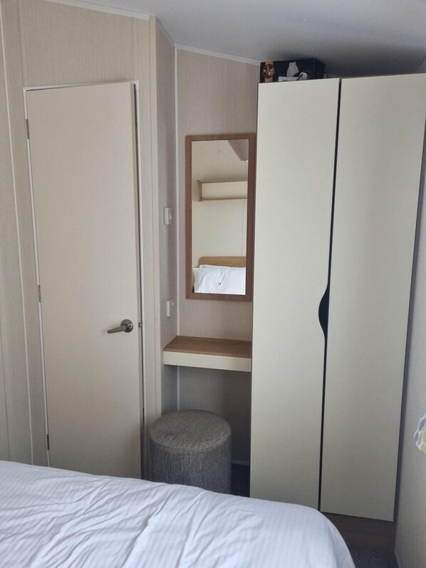 A bedroom with a bed, a wardrobe, a small stool, and a mirror. - 3 Bedroom Caravan, Sleeps 8, at Parkdean Newquay Holiday Park (Newquay)