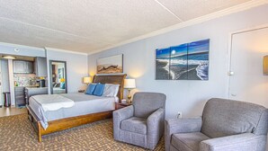 A hotel room with a bed, two armchairs, a painting of a beach, and a kitchenette. - Sand Dunes 2834 (Myrtle Beach)
