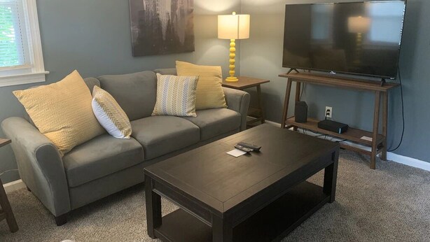 Un salon avec un canapé gris, des coussins jaunes, une table basse en bois, un téléviseur posé sur un meuble et une lampe sur une table d’appoint.