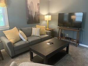 A living room with a grey sofa, yellow cushions, a wooden coffee table, a TV on a stand, and a lamp on a side table. - Newly Remodeled with deck. Close to town center (Wallingford)