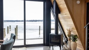 A modern interior with a large window overlooking a frozen body of water, a wooden staircase, and a black wood-burning stove. - Lakewood Lux Property - Saugatuck Harbor Luxury A-Frame, View of Downtown (Saugatuck)