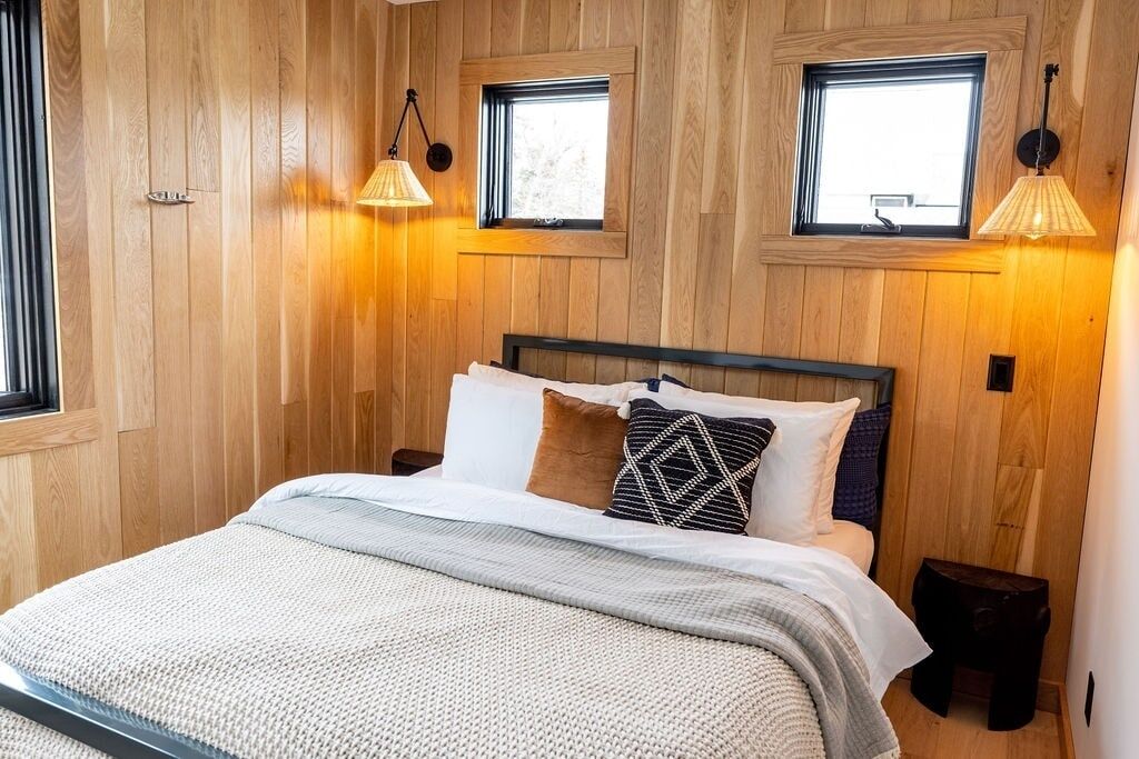 Una habitación con paneles de madera, una cama, dos ventanas y dos lámparas fijadas a la pared.