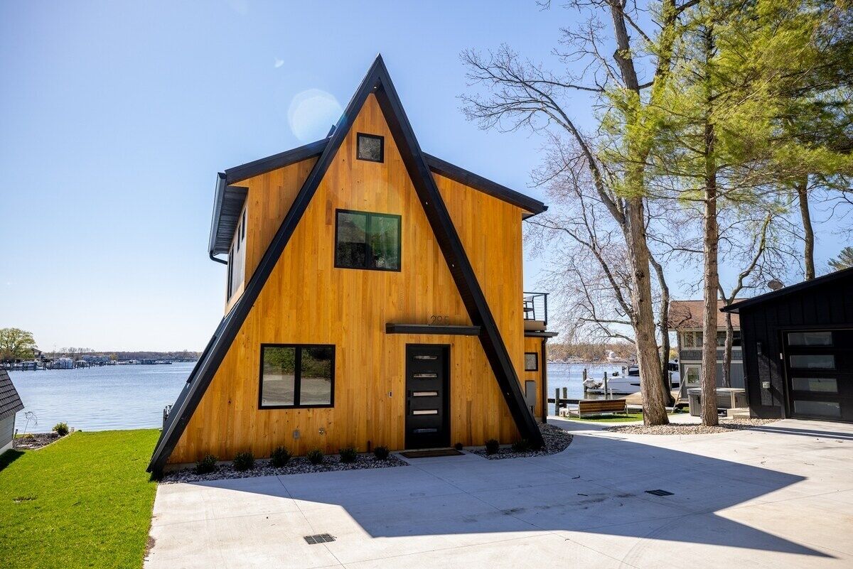 Une maison moderne en bois, dotée d’un toit triangulaire, d’une porte noire et de fenêtres, située au bord d’un plan d’eau.