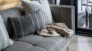 A modern interior with a wooden sloped ceiling, a grey sofa with patterned cushions, and a view of a waterfront through large windows. - Lakewood Lux Property - Saugatuck Harbor Luxury A-Frame, View of Downtown (Saugatuck)