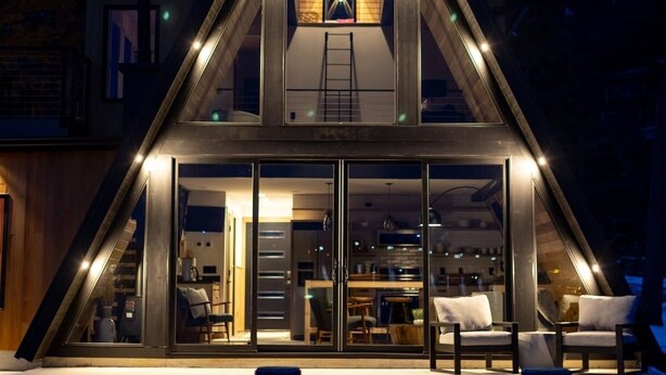 A modern, triangular-shaped building with a glass facade, a visible interior with seating and a kitchen area, and a ladder leading to an upper level. - Lakewood Lux Property - Saugatuck Harbor Luxury A-Frame, View of Downtown (Saugatuck)