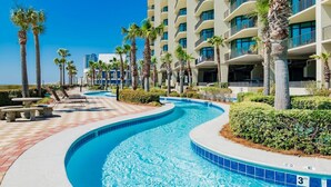 A swimming pool with a curved design, surrounded by palm trees and a multi-story hotel building. - Phoenix West 2107 | Orange Beach, AL (Orange Beach)