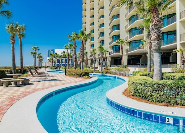 A swimming pool with a curved design, surrounded by palm trees and a multi-story hotel building.