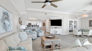A spacious living room with a ceiling fan, a large TV, and a dining area with white chairs. - Phoenix West 2107 | Orange Beach, AL (Orange Beach)