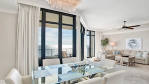 A modern living room with a glass dining table, white sofas, and a large window offering a view of the outdoors. - Phoenix West 2107 | Orange Beach, AL (Orange Beach)